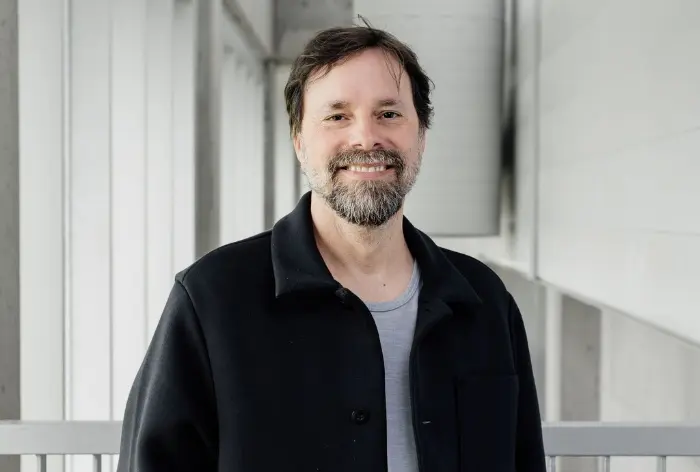 Patrice Aubertin, un homme barbu et moustachu, vêtu d'une veste noire sur une chemise grise, sourit dans un couloir moderne et lumineux aux murs et fenêtres blancs.