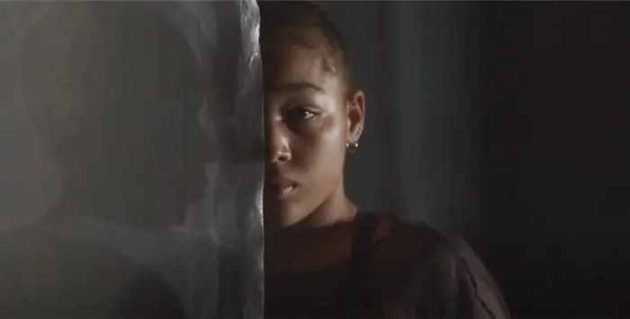 Une personne se tient partiellement cachée derrière un rideau transparent au festival du cirque actuel d'Auch, avec une moitié de son visage dans l'ombre et l'autre dans une lumière douce, créant une atmosphère sombre et contemplative.