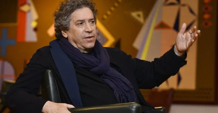 Un homme aux cheveux bouclés, vêtu d'un manteau et d'une écharpe sombres, est assis sur une chaise et fait un geste d'une main levée. L'arrière-plan présente un art géométrique abstrait aux couleurs chaudes, évoquant le flair théâtral du style visionnaire de Franco Dragone.