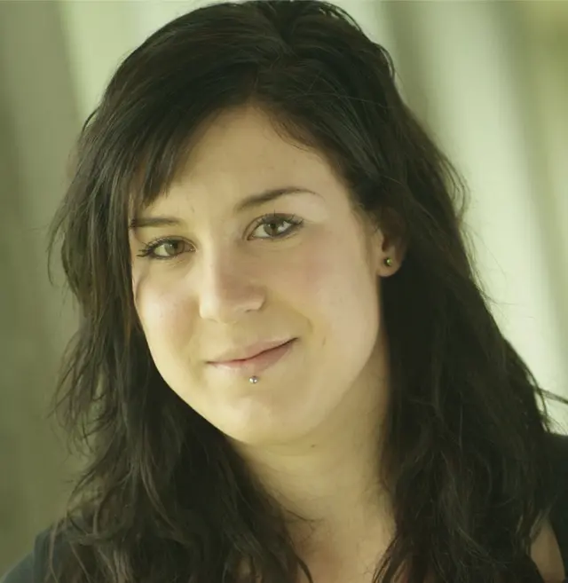 Valérie Côté, une femme aux longs cheveux noirs et à la peau claire, sourit doucement à la caméra. Elle a un petit piercing sous la lèvre inférieure et porte un haut noir. L'arrière-plan est légèrement flou, avec des tons verts et beiges.