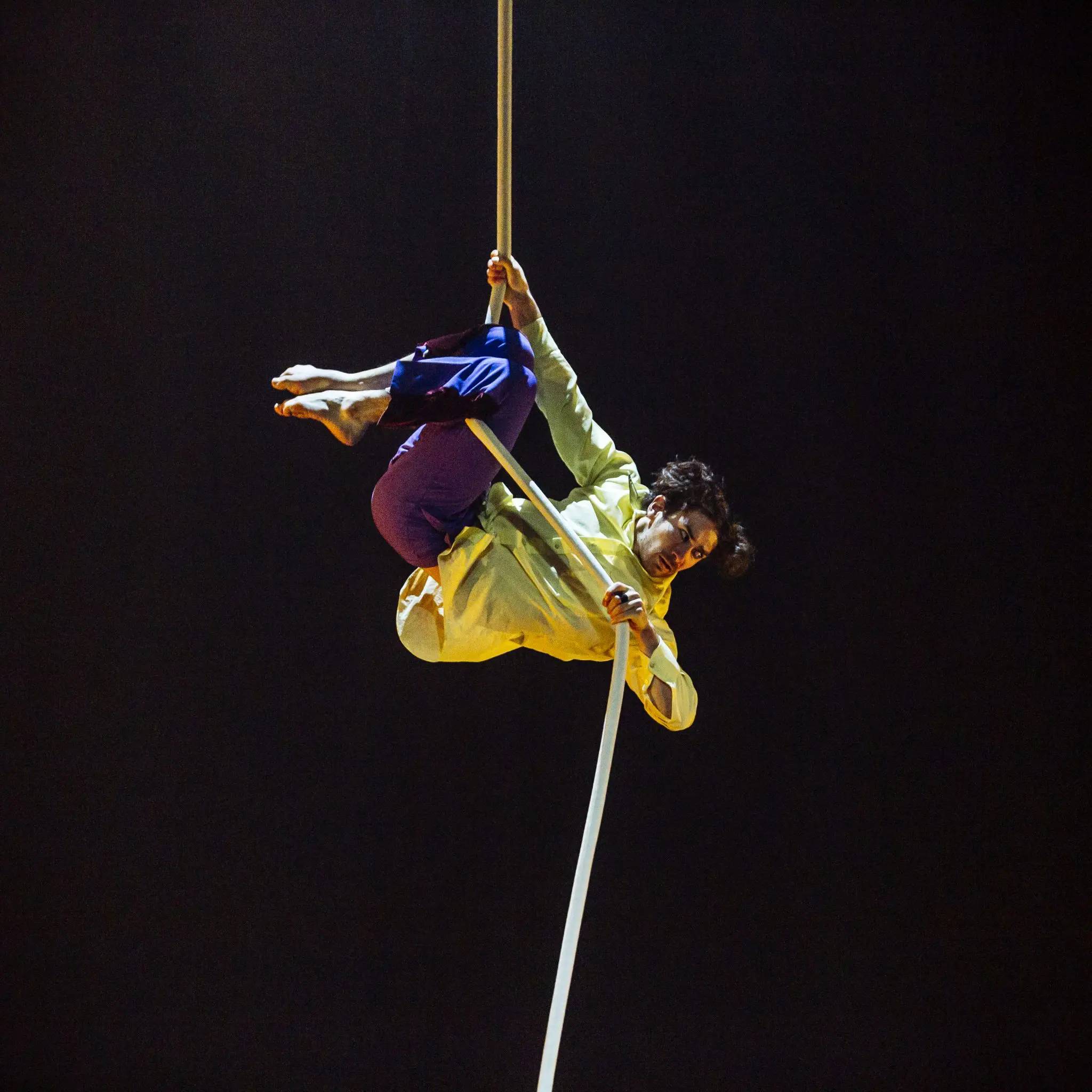 L'artiste aérien de la Revue Circus Arts est suspendu à l'envers dans des vêtements jaunes et violets, agrippant une longue corde blanche avec ses deux mains et ses deux jambes sur un fond sombre.