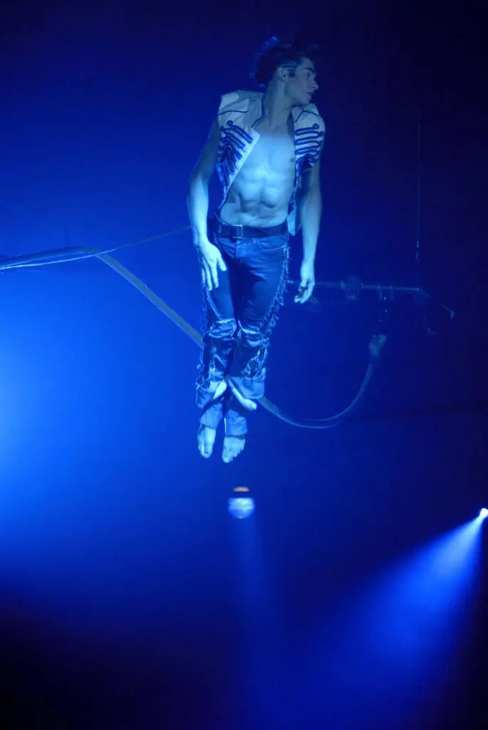 Un acrobate masculin à la carrure musclée évolue dans les airs sous un éclairage de scène bleu, vêtu d'un gilet et d'un pantalon ouverts. Pierre-Luc Houde apparaît suspendu à des fils, les pieds pointés vers le bas et la tête tournée sur le côté.