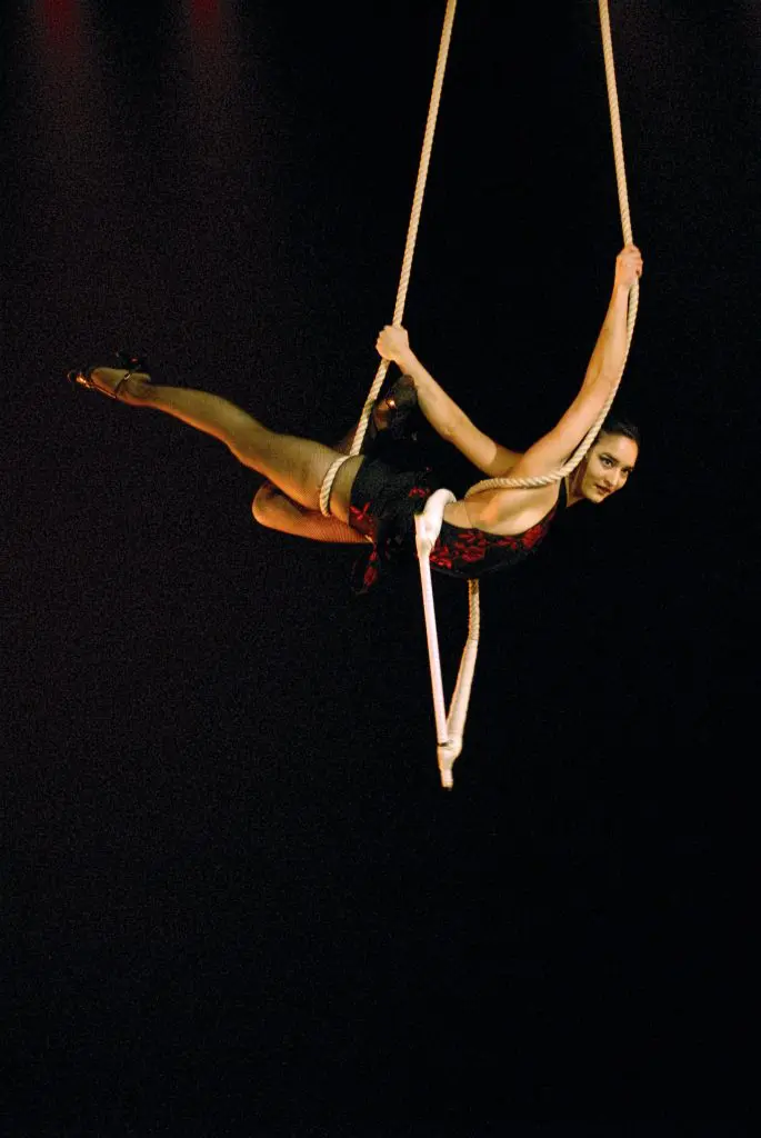 Marianella Michaud, acrobate féminine en costume noir et rouge, effectue un grand écart sur un trapèze, suspendue dans les airs sur un fond sombre, un bras et une jambe gracieusement tendus.