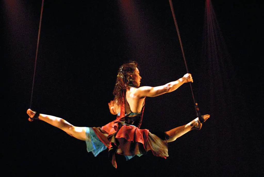 L'artiste aérienne Kylee Moats Maupoux, vêtue d'une robe colorée, fait un grand écart en étant suspendue à des cordes sous un éclairage dramatique sur un fond sombre.