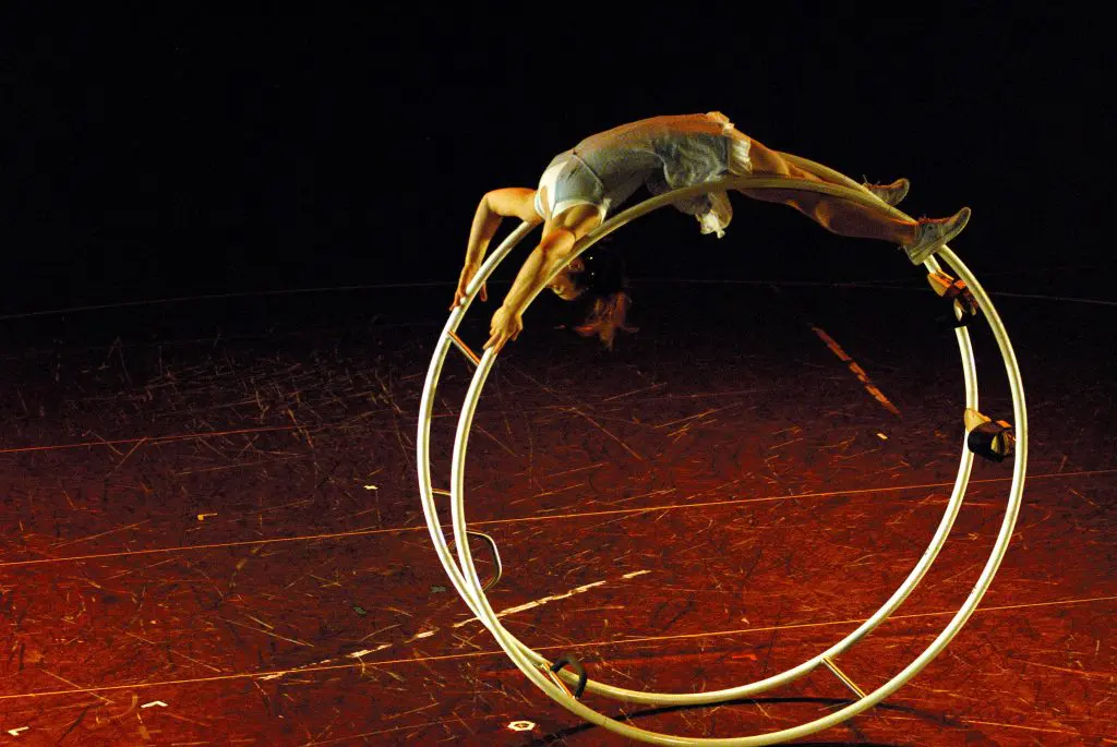 Une interprète vêtue d'une robe légère, rappelant l'élégance de Marie-Claude Roulez, se tient en équilibre la tête en bas sur une grande roue métallique sur scène, arquant gracieusement le dos tout en saisissant la roue avec ses pieds et ses mains sous un éclairage dramatique.