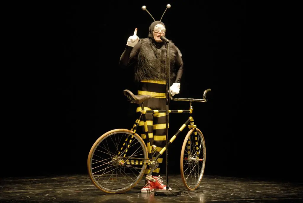 Une personne vêtue d'un costume d'abeille se tient derrière un vélo rayé jaune et noir sur une scène sombre, parlant dans un microphone et levant un doigt, portant des gants blancs et des chaussures rouges - un acte inspiré par Rubén Martin Urdiales.