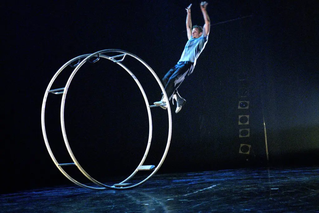 Arno Wauters effectue une acrobatie, suspendu à une grande roue métallique sur une scène sombre, avec un éclairage dramatique mettant en valeur l'artiste et l'équipement.