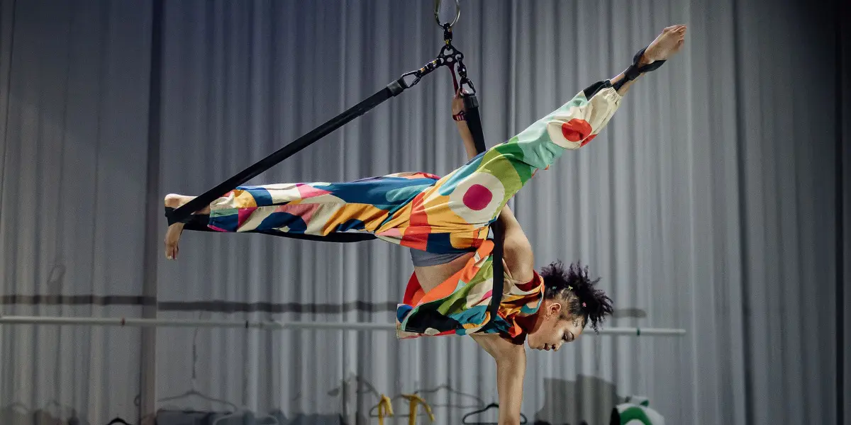 Un artiste de cirque vêtu d'un costume coloré à motifs est suspendu horizontalement par un harnais et des sangles, un bras tendu. Derrière eux, plusieurs costumes vibrants sont suspendus sur une scène éclairée par des rideaux, capturant le dynamisme artistique de Zoe Schubert.