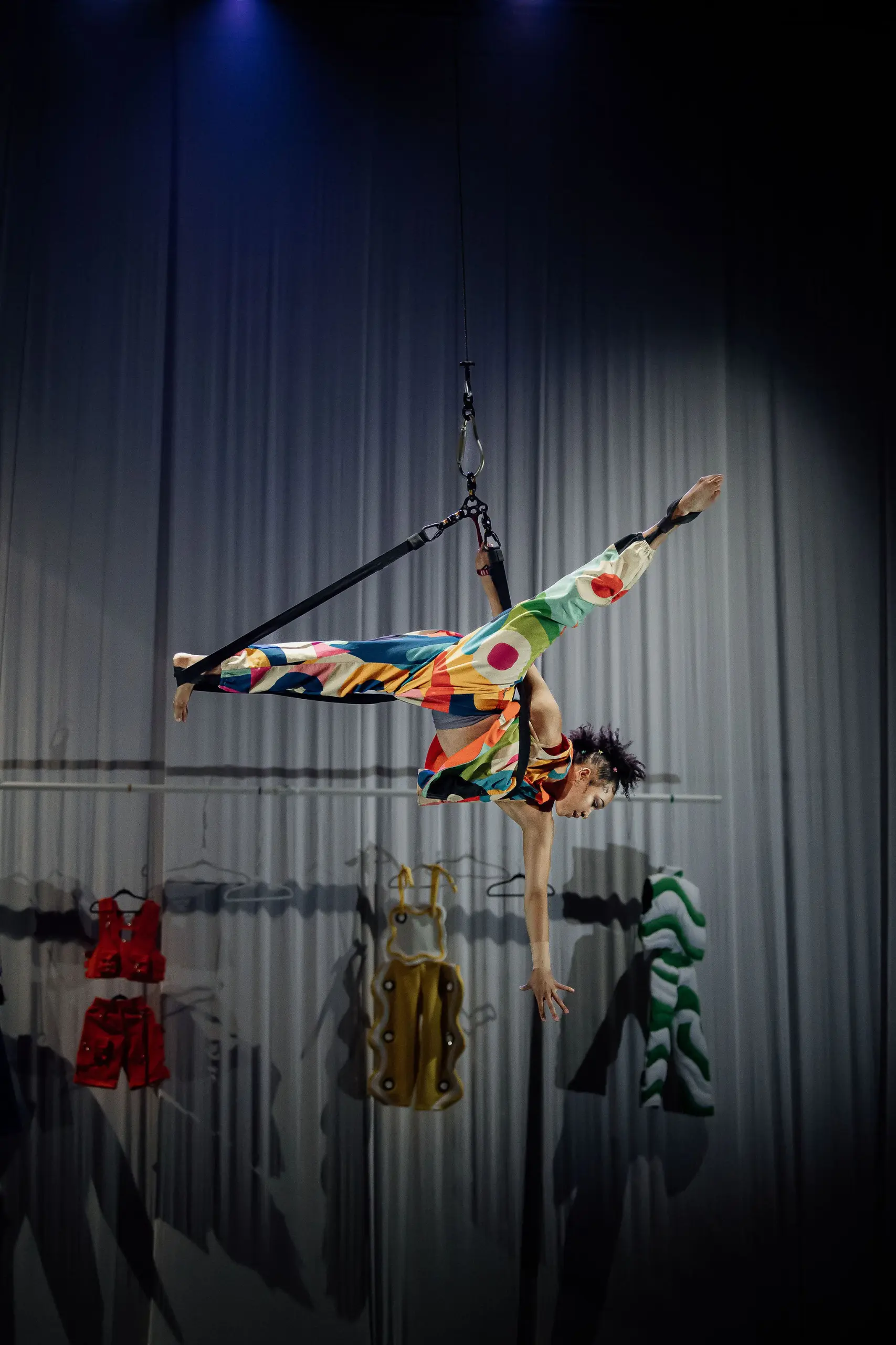 Un artiste de cirque vêtu d'un costume coloré à motifs est suspendu horizontalement par un harnais et des sangles, un bras tendu. Derrière eux, plusieurs costumes vibrants sont suspendus sur une scène éclairée par des rideaux, capturant le dynamisme artistique de Zoe Schubert.