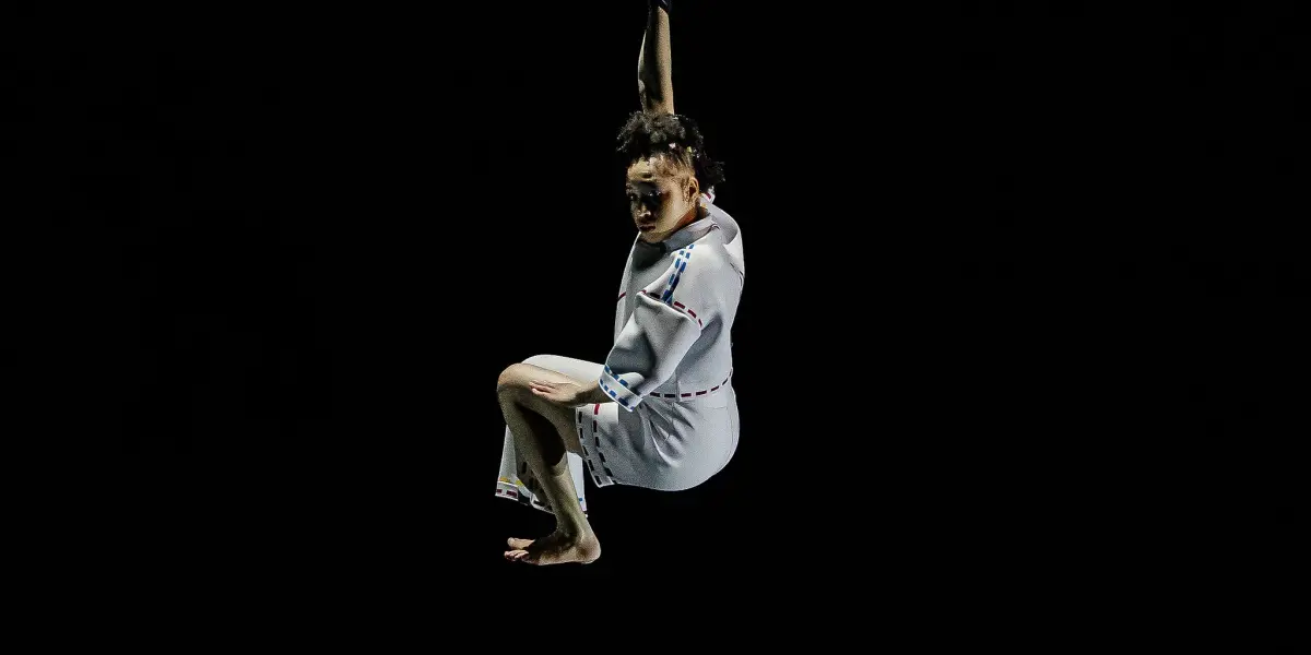 Zoe Schubert, vêtue d'une tenue blanche, est suspendue dans les airs sur un fond entièrement noir, tenant une corde au-dessus de sa tête et regardant vers le bas avec un genou plié.