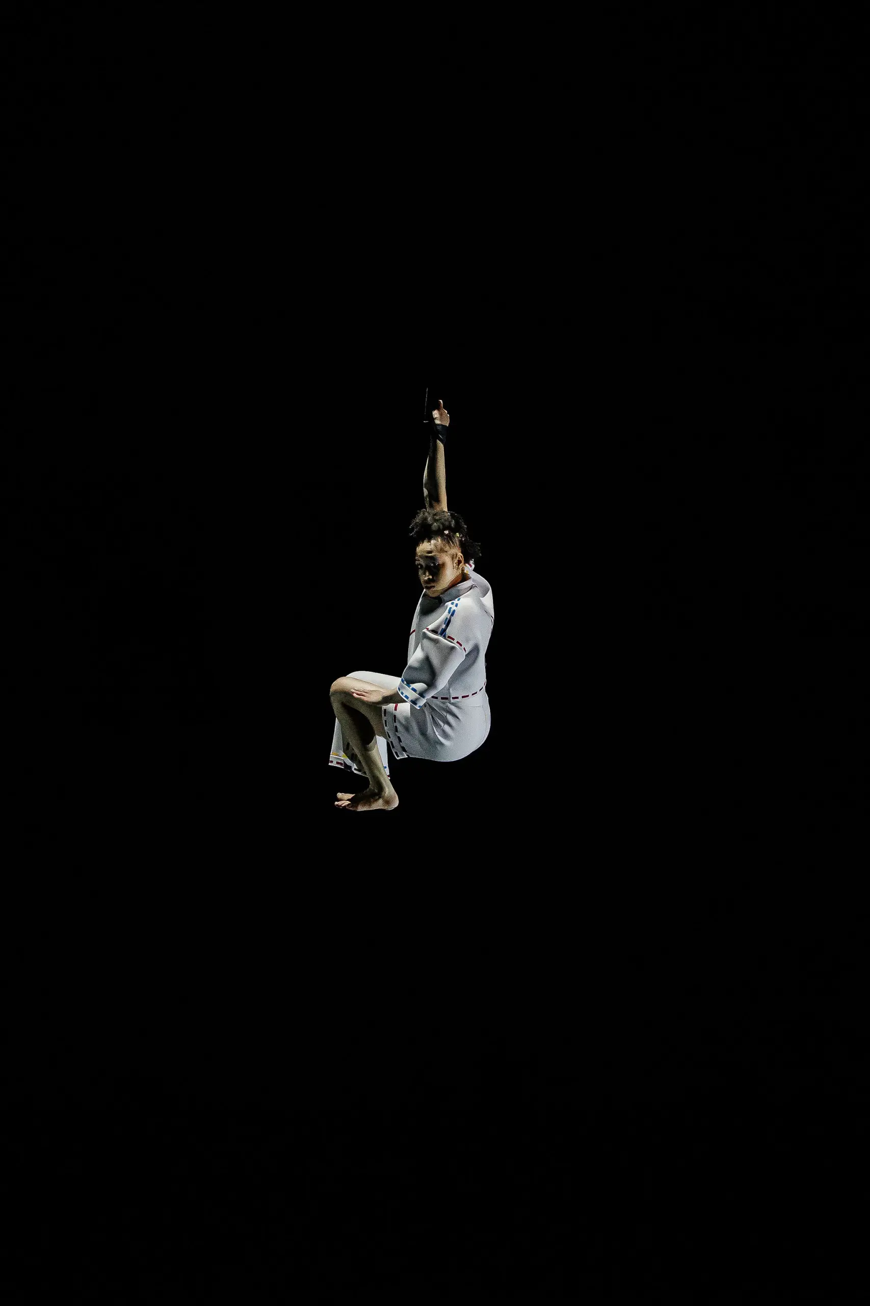 Zoe Schubert, vêtue d'une tenue blanche, est suspendue dans les airs sur un fond entièrement noir, tenant une corde au-dessus de sa tête et regardant vers le bas avec un genou plié.