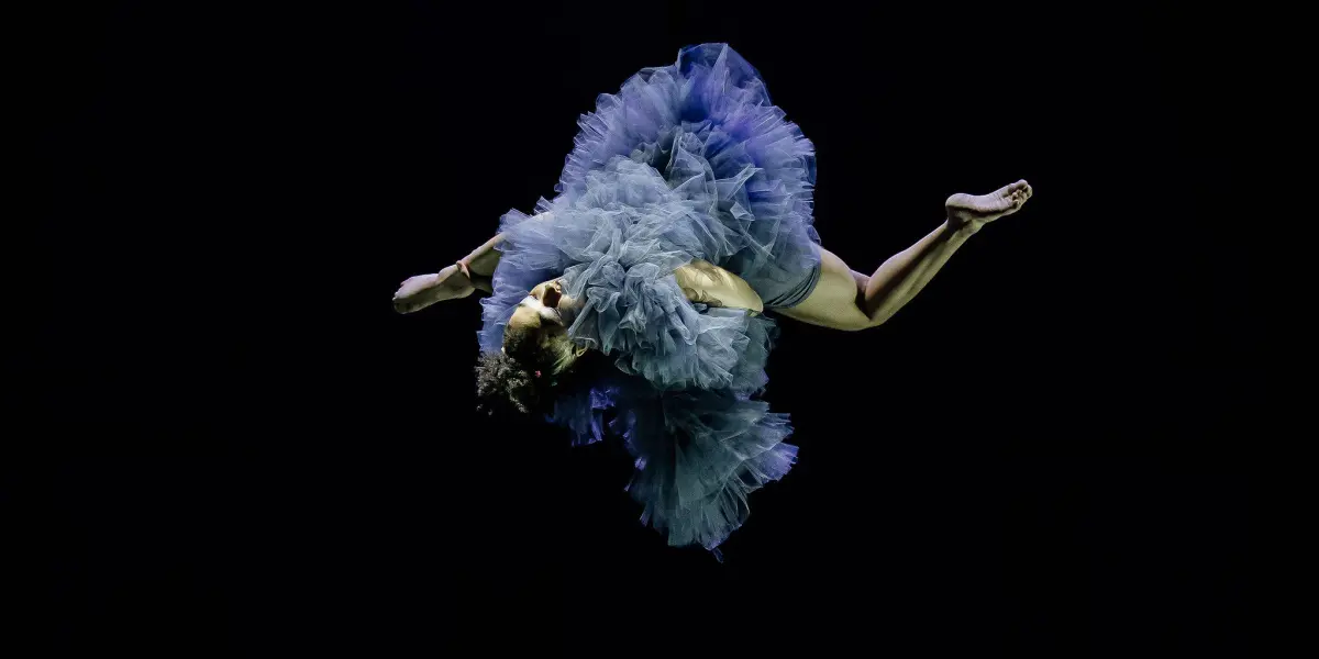 L'artiste aérienne Zoe Schubert est suspendue à l'envers dans les airs, vêtue d'un volumineux costume bleu à volants sur fond noir, les bras et les jambes tendus vers l'extérieur.