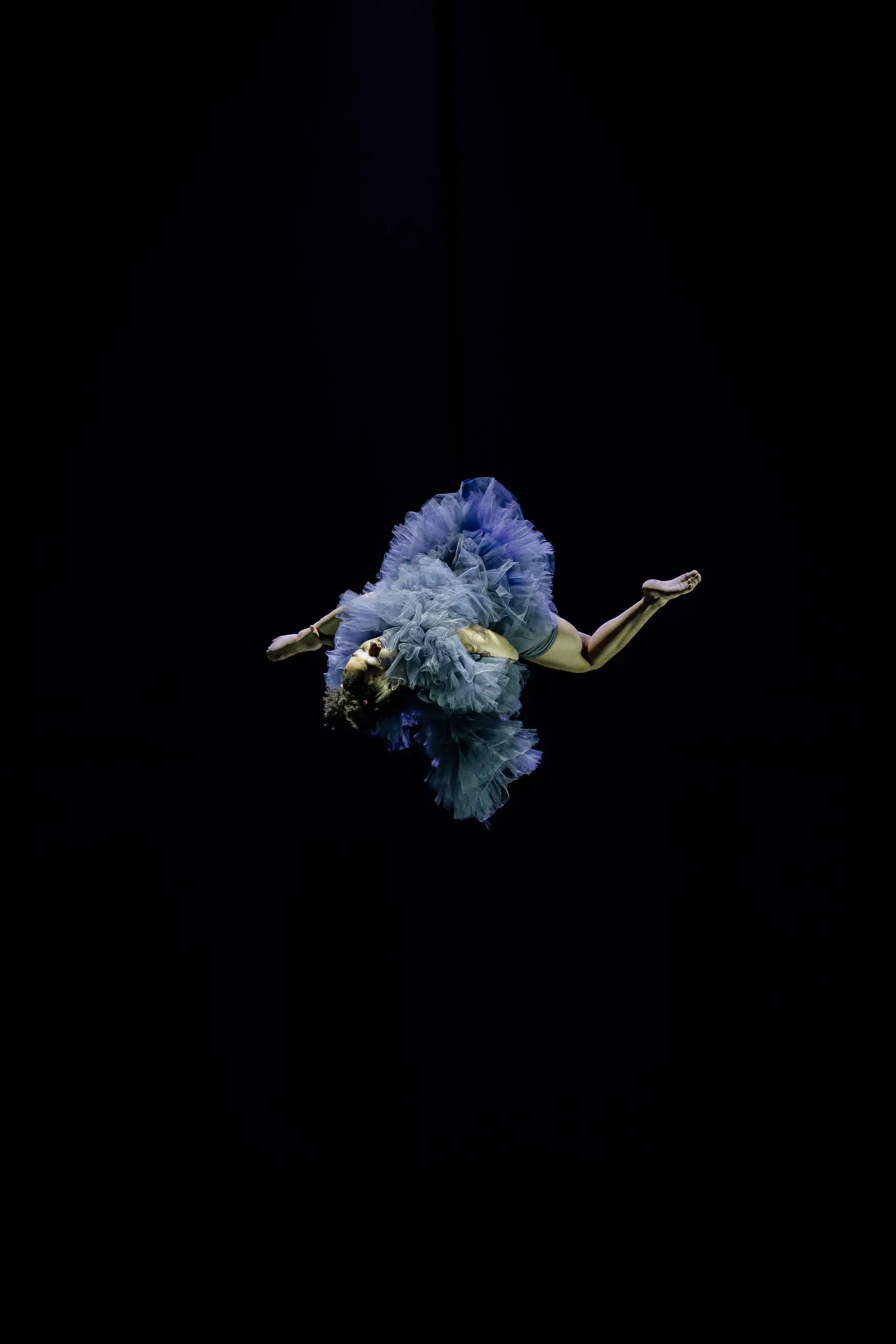 L'artiste aérienne Zoe Schubert est suspendue à l'envers dans les airs, vêtue d'un volumineux costume bleu à volants sur fond noir, les bras et les jambes tendus vers l'extérieur.