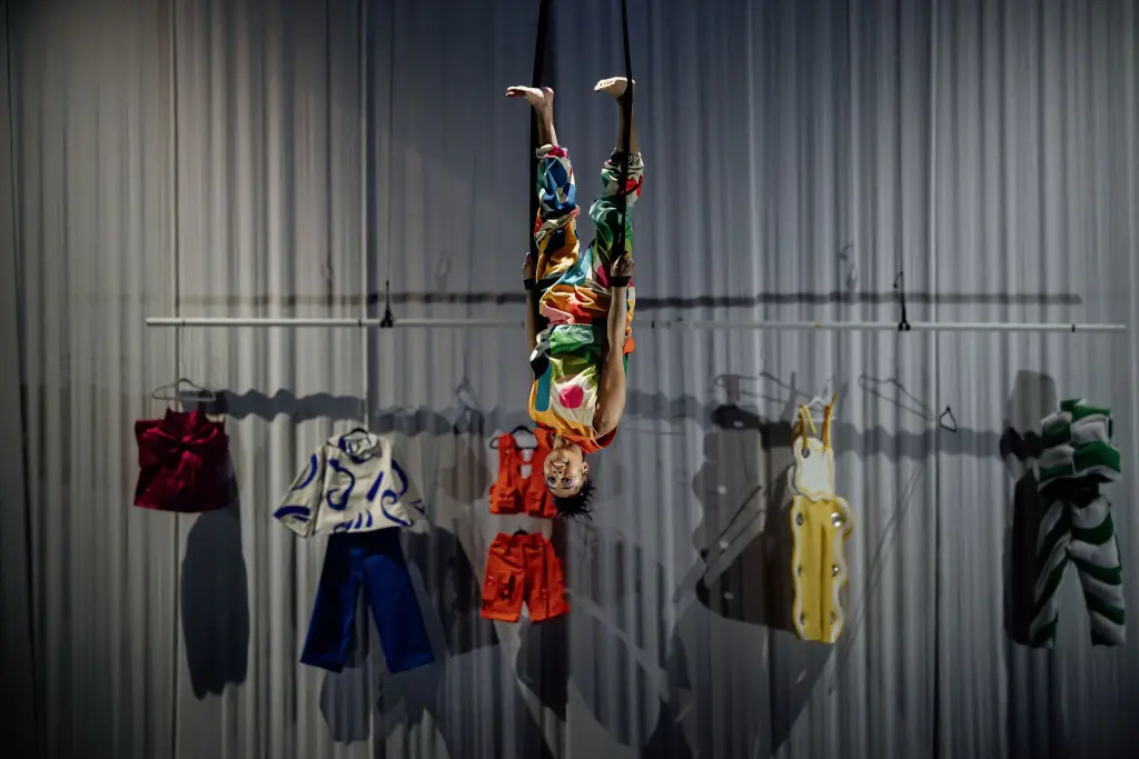 Une personne vêtue de vêtements colorés est suspendue à un trapèze, la tête en bas, tandis qu'un éclairage dramatique projette des ombres sur le rideau à l'arrière. Parmi les tenues vibrantes sur les cintres, une pièce de Zoe Schubert se distingue dans cette scène animée.