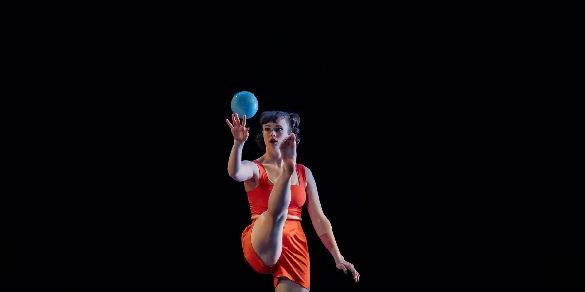 Isabella Majzun, en tenue orange, se tient en équilibre sur une jambe tout en lançant une balle bleue en l'air, sur un fond sombre.