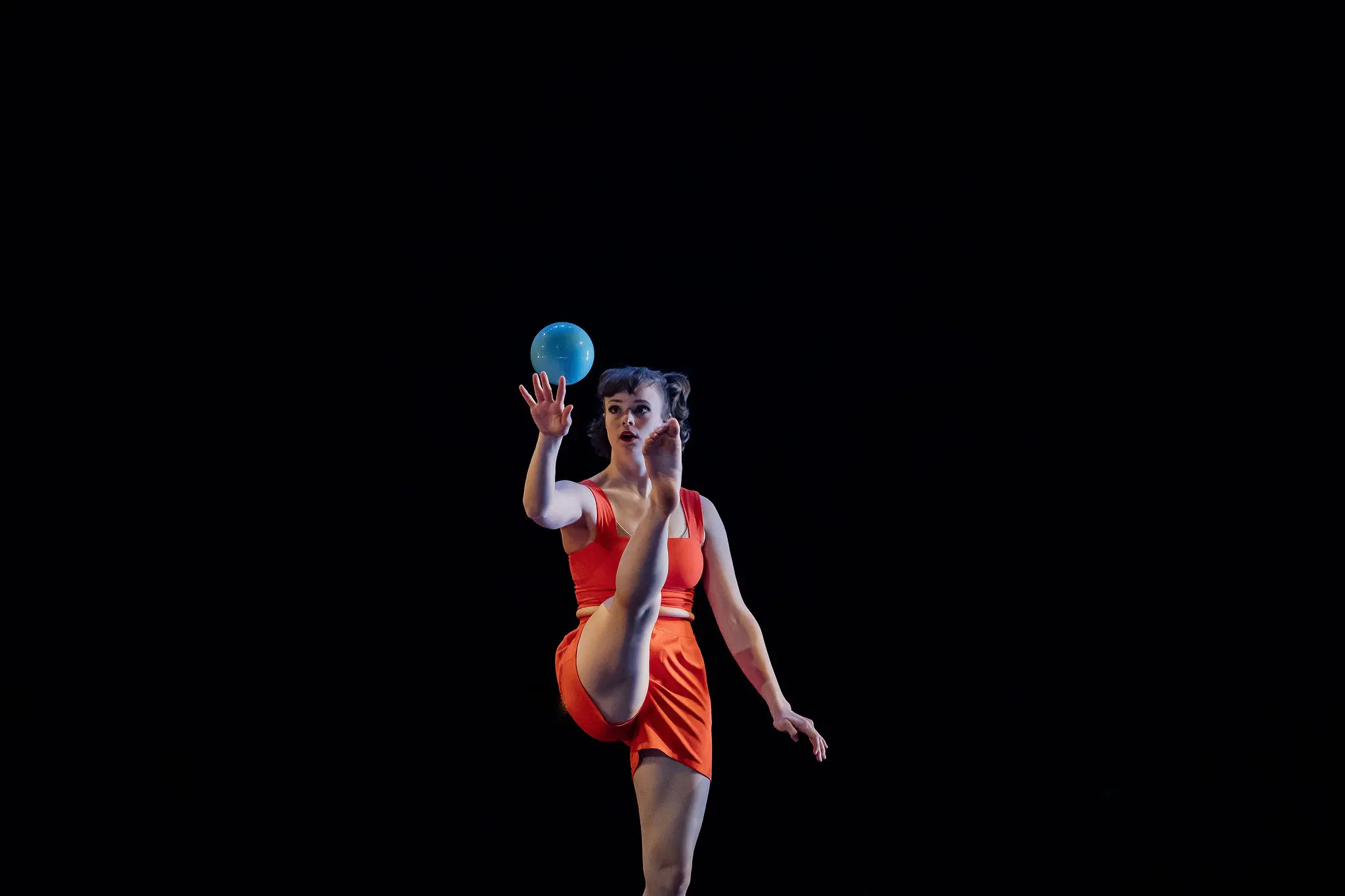Isabella Majzun, en tenue orange, se tient en équilibre sur une jambe tout en lançant une balle bleue en l'air, sur un fond sombre.