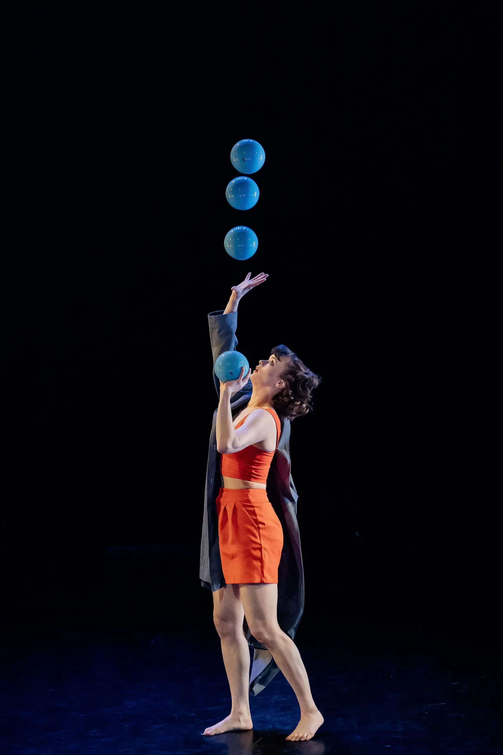 Isabella Majzun, pieds nus dans une tenue orange et une robe sombre, jongle habilement avec cinq balles bleues sur une scène sombre.