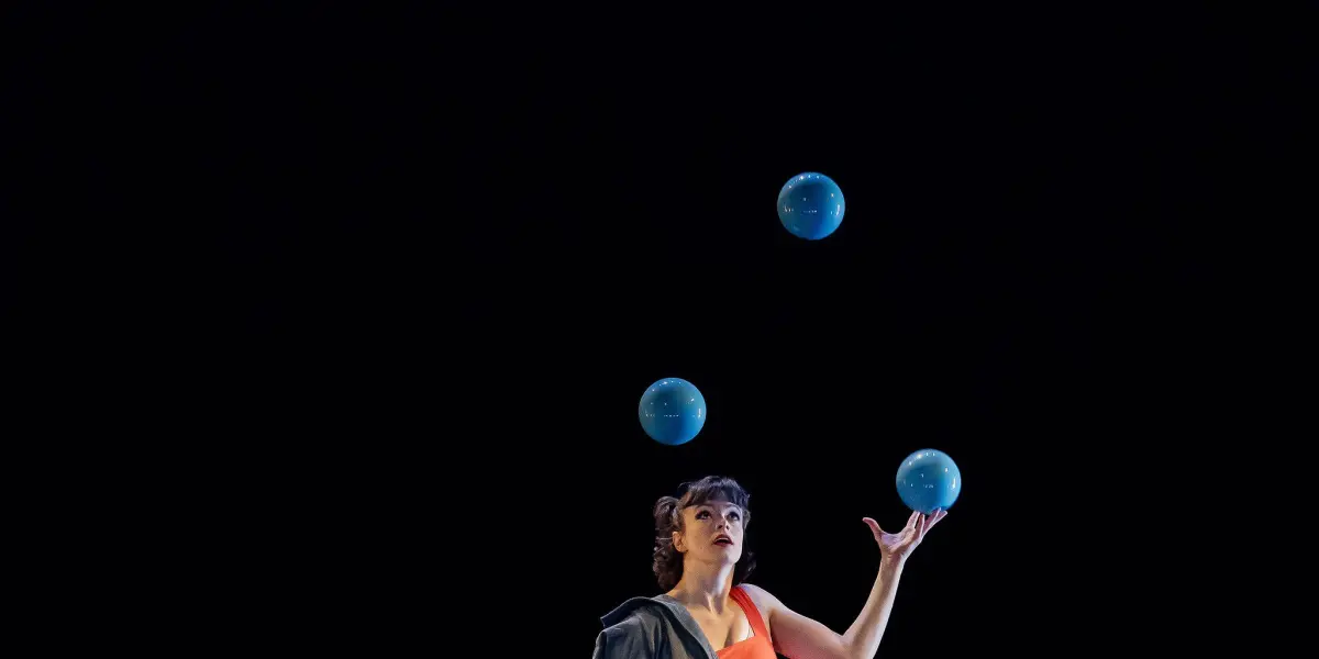 Isabella Majzun, vêtue d'une tenue orange et drapée dans un manteau gris, jongle avec quatre balles bleues sous un projecteur sur fond noir.