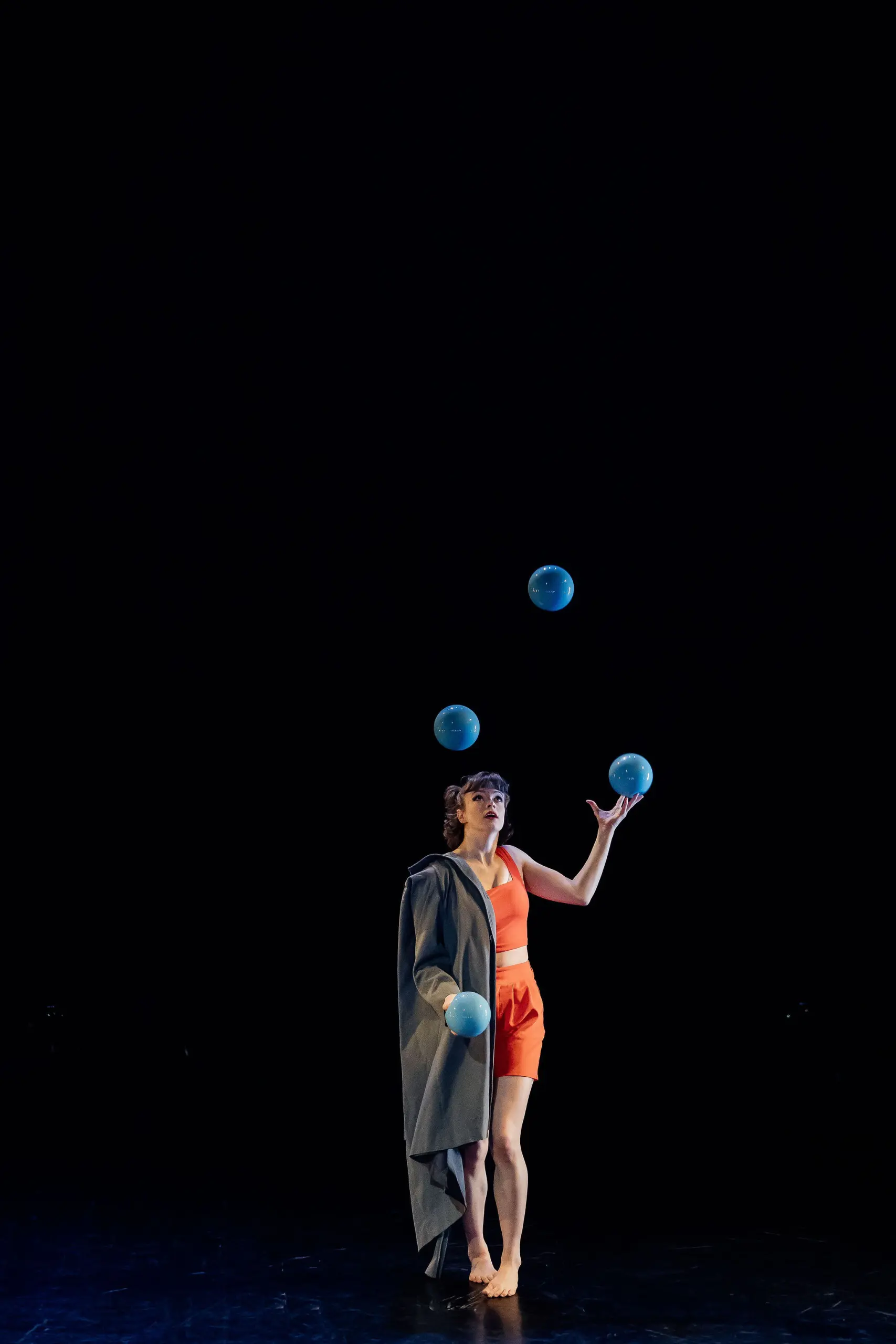 Isabella Majzun, vêtue d'une tenue orange et drapée dans un manteau gris, jongle avec quatre balles bleues sous un projecteur sur fond noir.