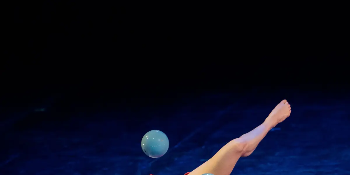 Isabella Majzun, vêtue d'orange, jongle avec quatre balles bleues, allongée sur le dos, sur une scène sombre. Une jambe levée et les bras tendus, elle s'exécute habilement sous les feux de la rampe.
