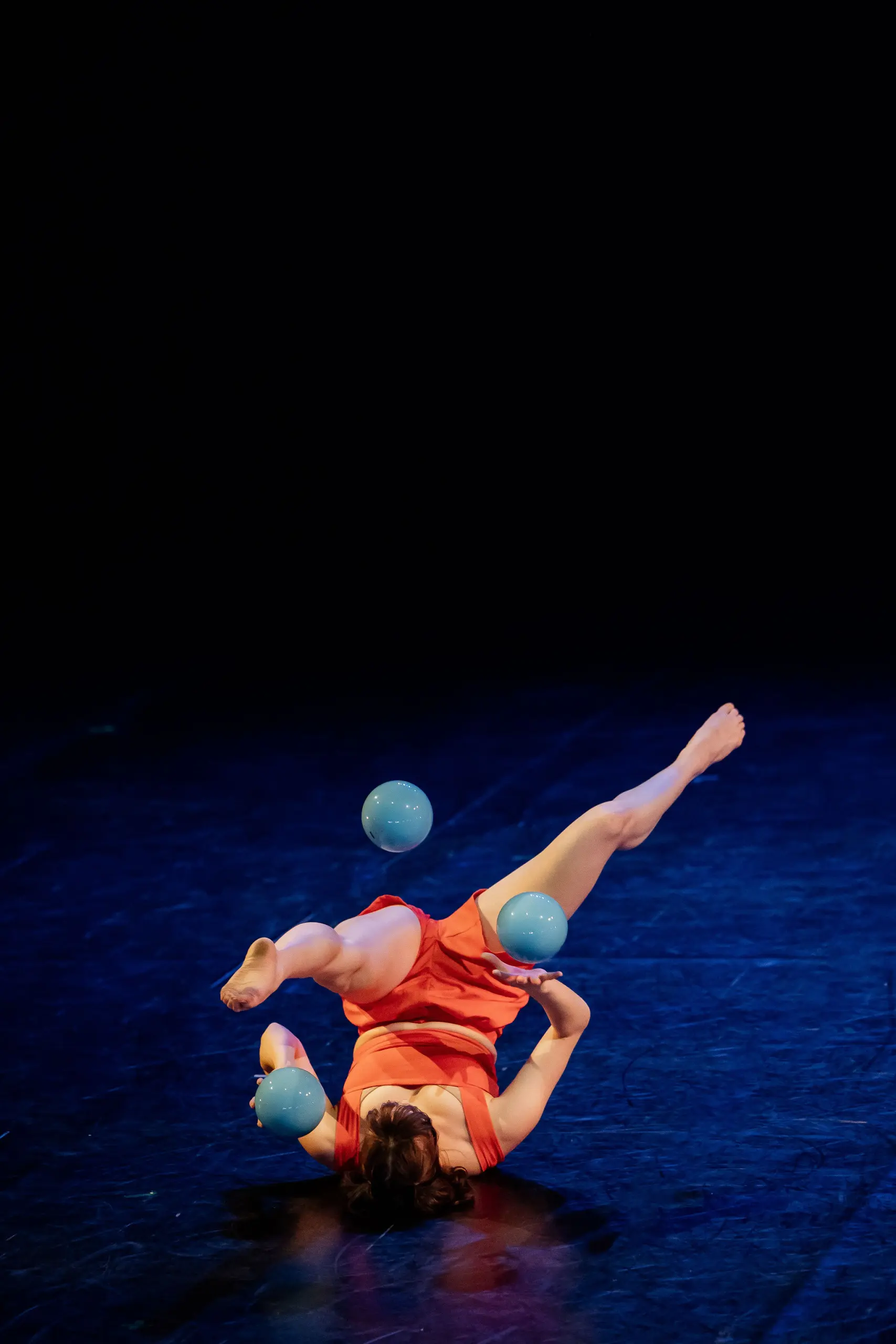 Isabella Majzun, vêtue d'orange, jongle avec quatre balles bleues, allongée sur le dos, sur une scène sombre. Une jambe levée et les bras tendus, elle s'exécute habilement sous les feux de la rampe.