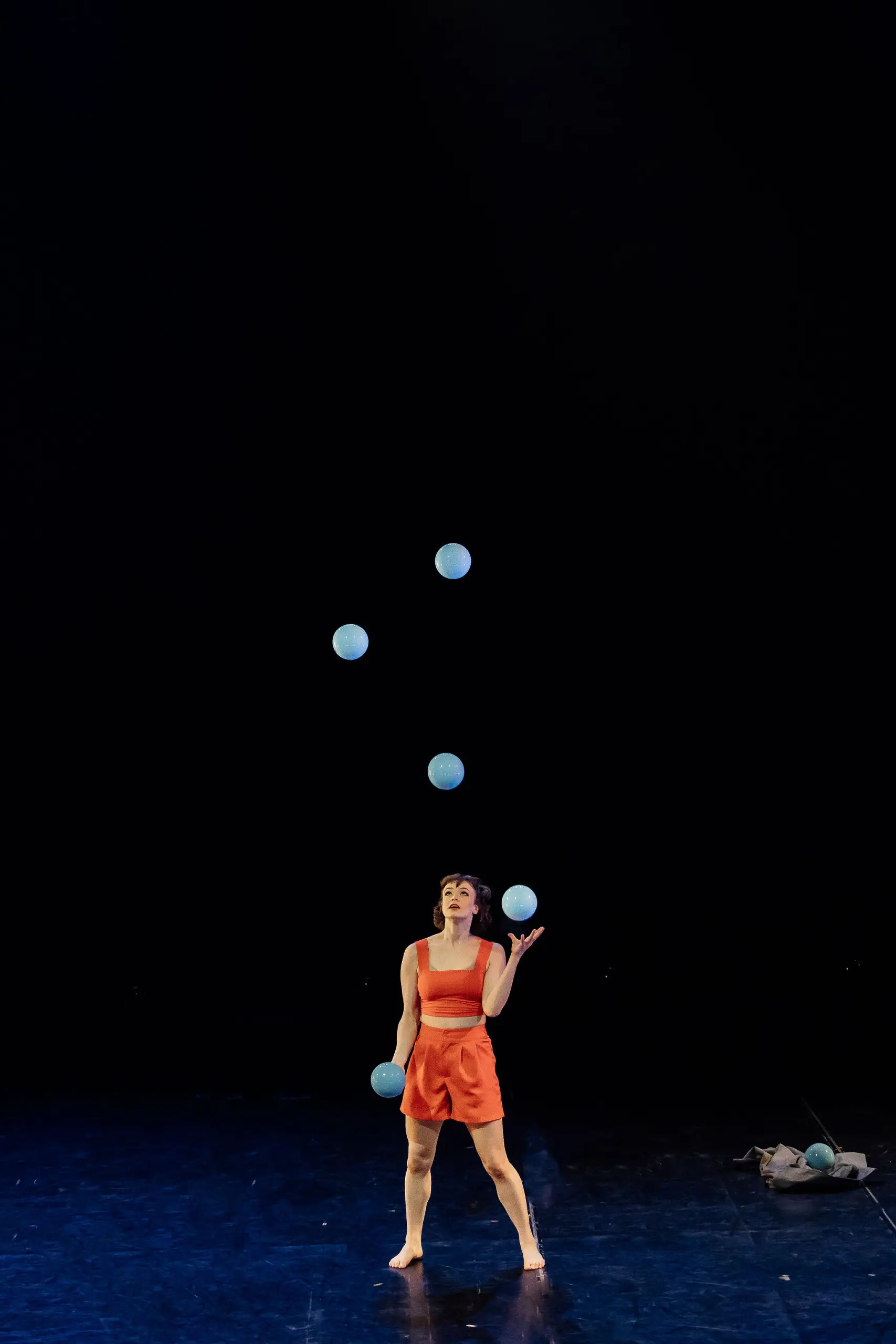 Isabella Majzun, vêtue d'orange, jongle avec cinq balles bleues sur une scène sombre, avec une petite pile d'objets sur le sol à proximité.