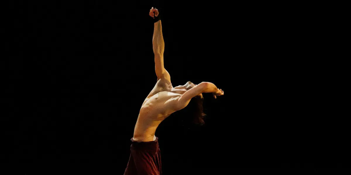 Un danseur contemporain, Iason Ioannidis, torse nu et pantalon rouge, prend une pose théâtrale sur une scène sombre, en arquant le dos et en étirant un bras vers le haut, sous l'éclairage d'un projecteur sur fond noir.