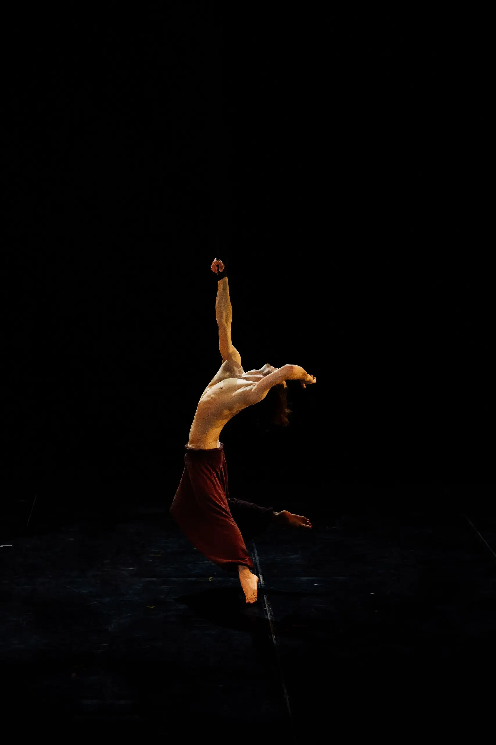 Un danseur contemporain, Iason Ioannidis, torse nu et pantalon rouge, prend une pose théâtrale sur une scène sombre, en arquant le dos et en étirant un bras vers le haut, sous l'éclairage d'un projecteur sur fond noir.