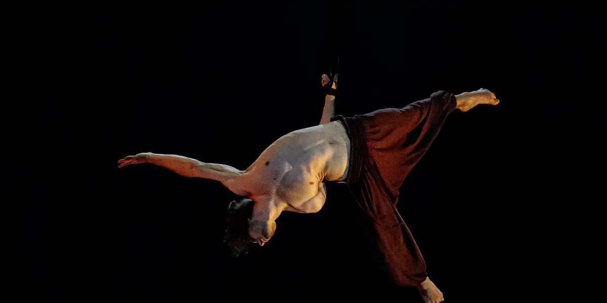 Un artiste aérien torse nu, Iason Ioannidis, vêtu d'un pantalon sombre ample, est suspendu à l'envers, bras et jambes tendus, par une sangle, sous un projecteur, sur un fond sombre.