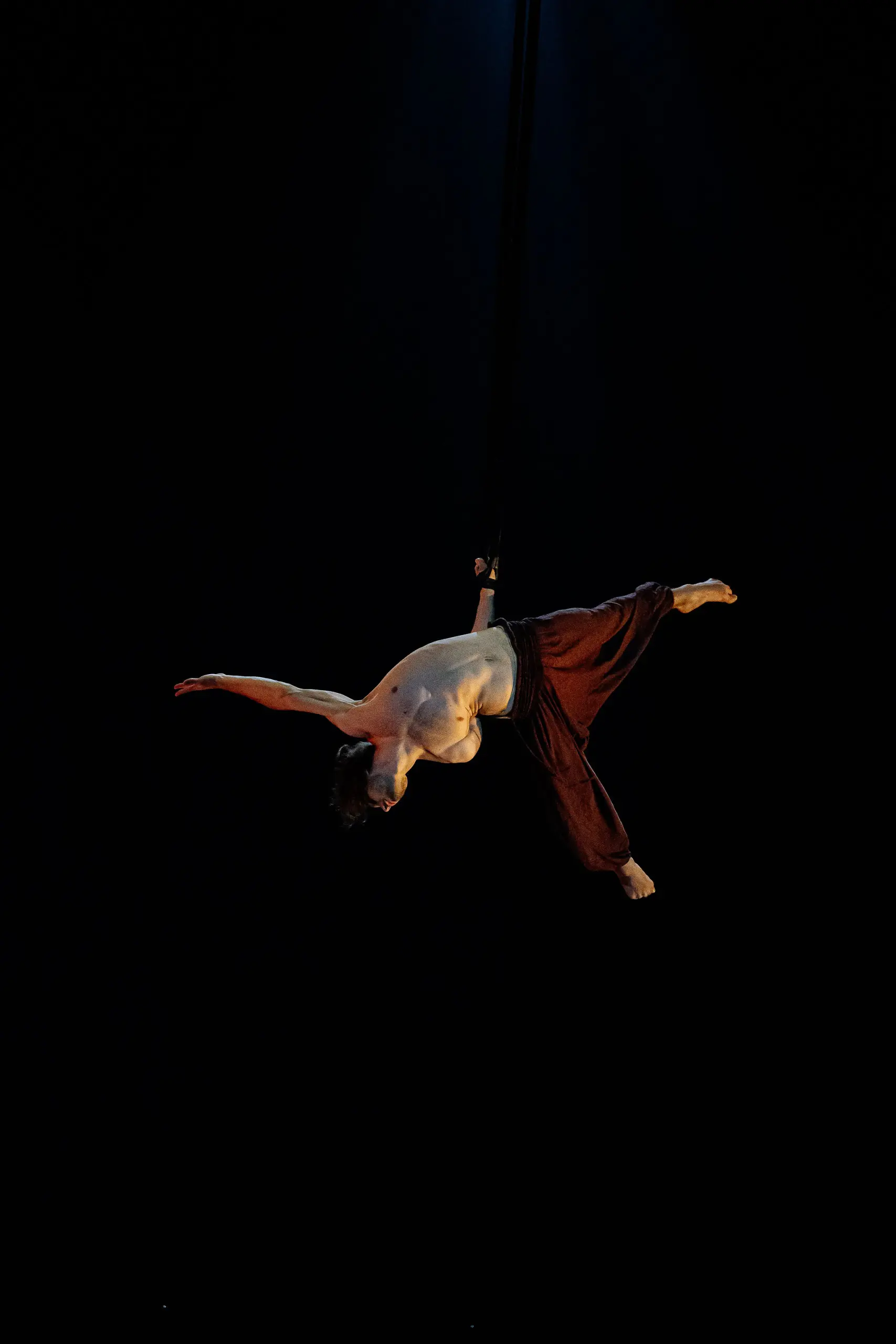 Un artiste aérien torse nu, Iason Ioannidis, vêtu d'un pantalon sombre ample, est suspendu à l'envers, bras et jambes tendus, par une sangle, sous un projecteur, sur un fond sombre.