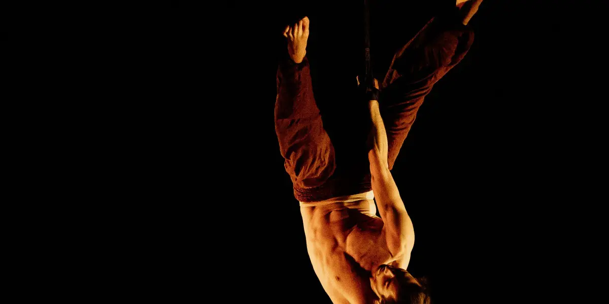 Un artiste aérien torse nu, Iason Ioannidis, est suspendu à l'envers en tenant une sangle d'une main sur un fond noir. Son bras et ses jambes sont étendus dans une pose dynamique, mise en valeur par un éclairage dramatique.