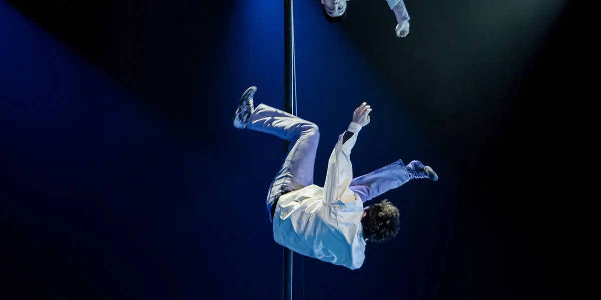 Deux acrobates vêtus de chemises blanches et de pantalons amples exécutent une acrobatie aérienne sur un mât vertical, suspendus la tête en bas sous un éclairage de scène bleu spectaculaire - un spectacle époustouflant de Victor Crepin.