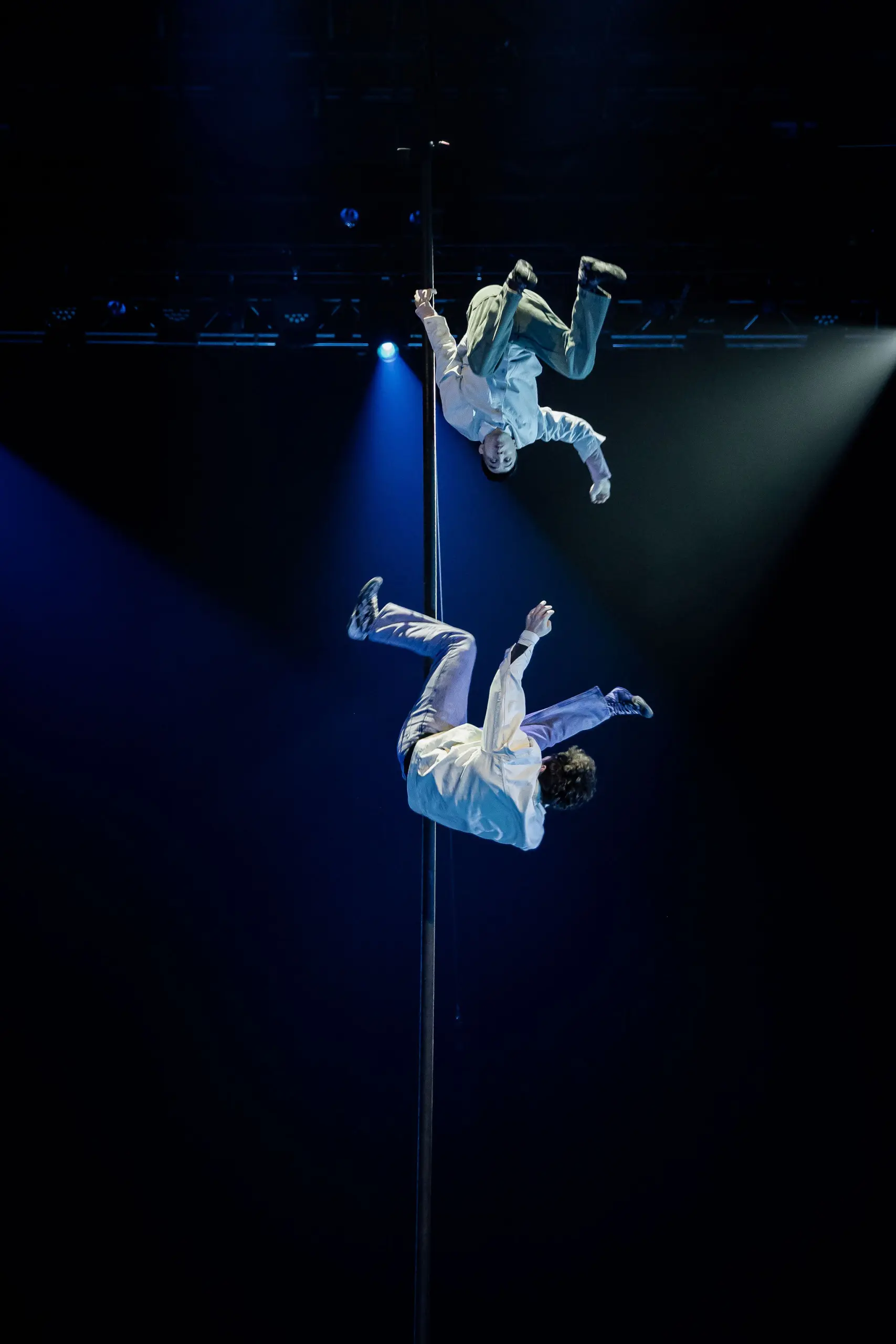 Deux acrobates vêtus de chemises blanches et de pantalons amples exécutent une acrobatie aérienne sur un mât vertical, suspendus la tête en bas sous un éclairage de scène bleu spectaculaire - un spectacle époustouflant de Victor Crepin.