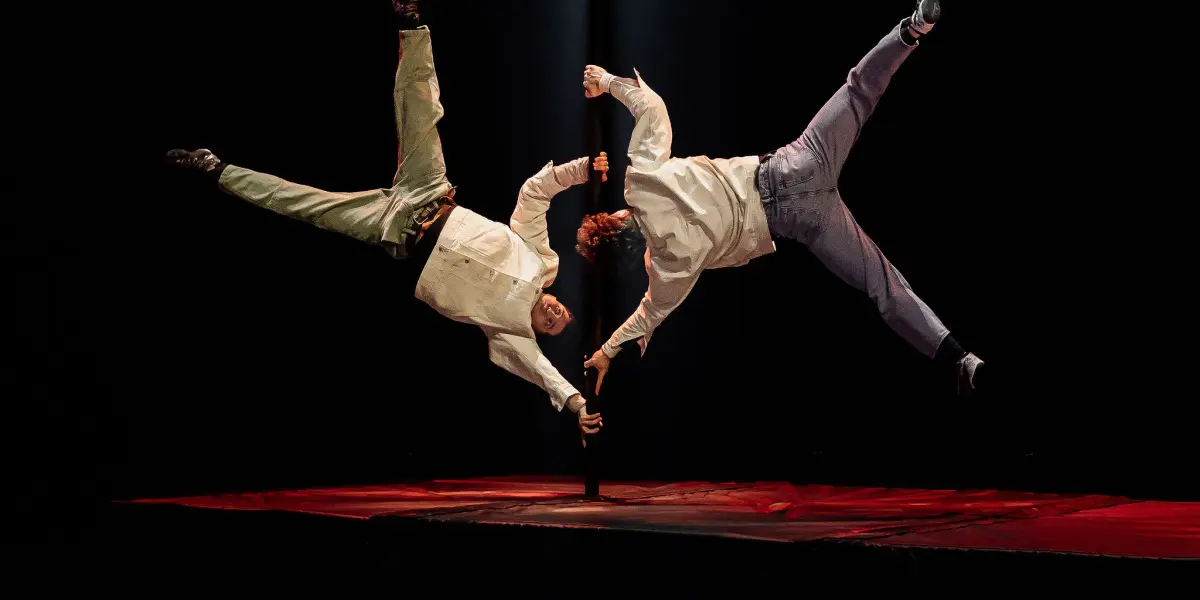 Deux acrobates, dont Victor Crépin, exécutent une acrobatie aérienne sur une scène sombre, se tenant d'une main alors qu'ils flottent à l'horizontale, éclairés par un projecteur dramatique venant d'en haut.