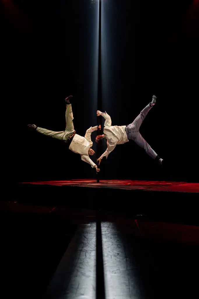 Deux acrobates, dont Victor Crépin, exécutent une acrobatie aérienne sur une scène sombre, se tenant d'une main alors qu'ils flottent à l'horizontale, éclairés par un projecteur dramatique venant d'en haut.