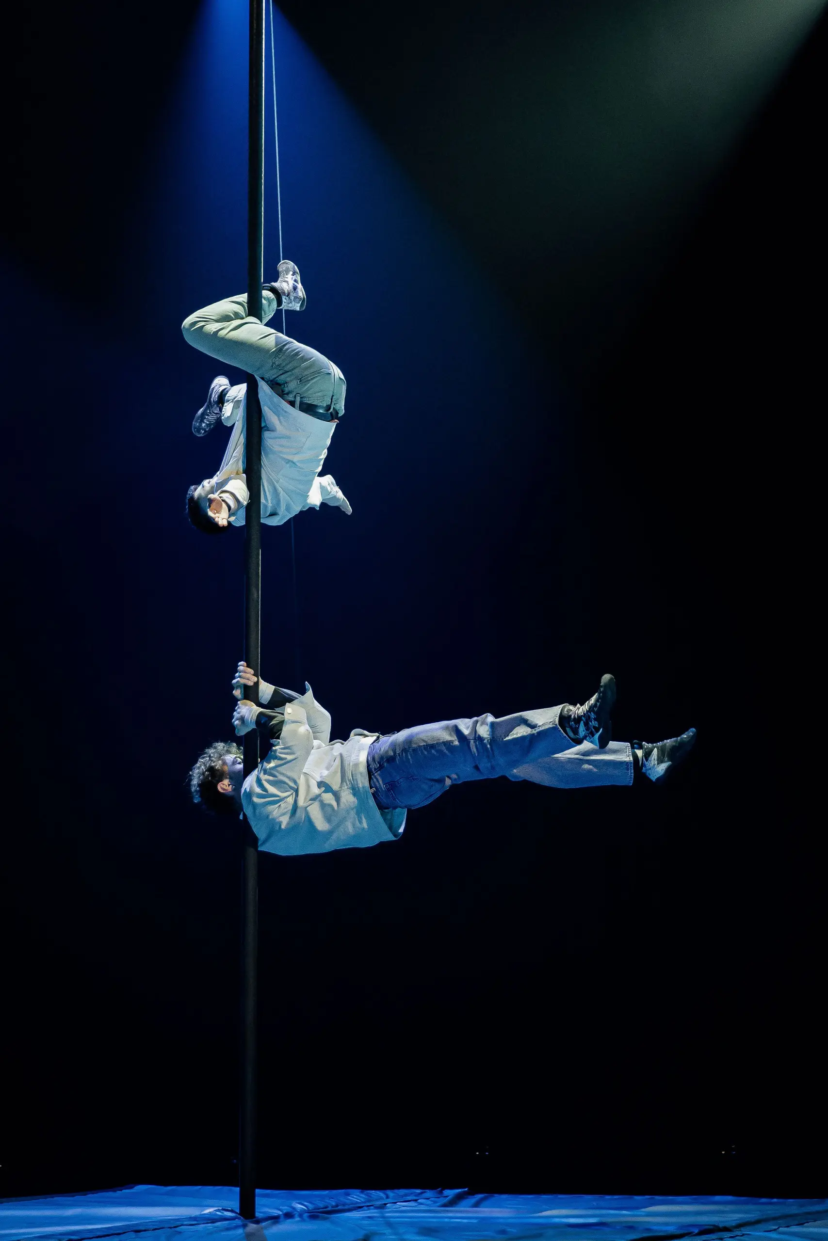 Deux acrobates en tenue blanche évoluent sur un mât vertical sous un éclairage scénique spectaculaire. L'un, Victor Crepin, est suspendu à l'envers près du sommet tandis que l'autre tient le mât à l'horizontale, les jambes étendues.