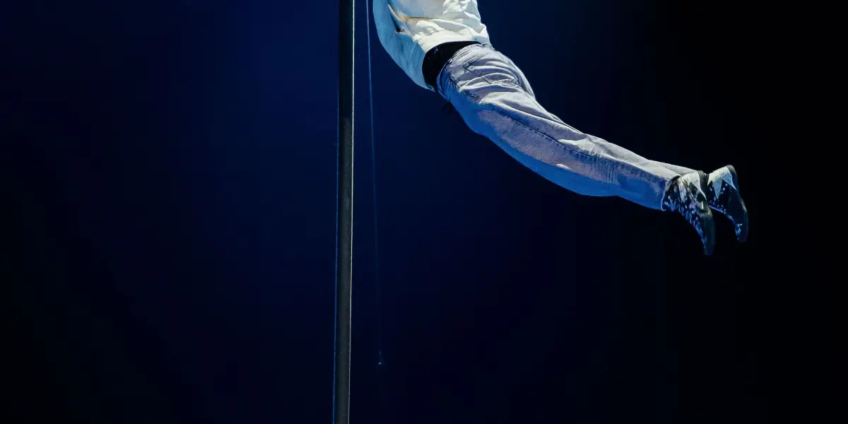 Deux acrobates, dont Victor Crépin, se produisent sur un poteau vertical sous un éclairage dramatique ; l'un est suspendu latéralement près du sommet, agrippé au poteau, tandis que l'autre est suspendu à l'envers plus bas sur le poteau, tous deux en tenue décontractée.