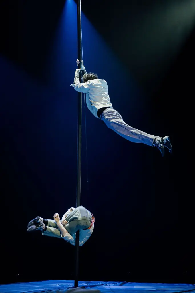 Deux acrobates, dont Victor Crépin, se produisent sur un poteau vertical sous un éclairage dramatique ; l'un est suspendu latéralement près du sommet, agrippé au poteau, tandis que l'autre est suspendu à l'envers plus bas sur le poteau, tous deux en tenue décontractée.