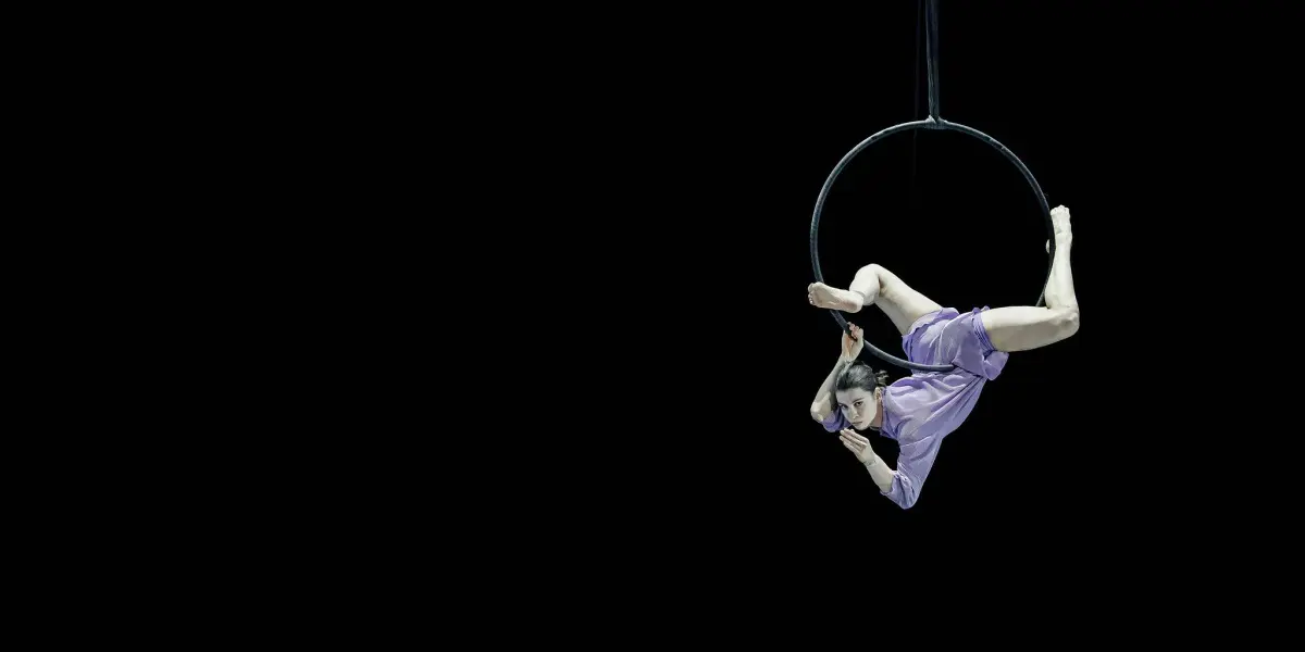 L'artiste aérienne Katarina Sobinkovicova, vêtue de violet, est suspendue à l'envers à un cerceau sur fond noir, faisant preuve d'une souplesse et d'un équilibre remarquables.
