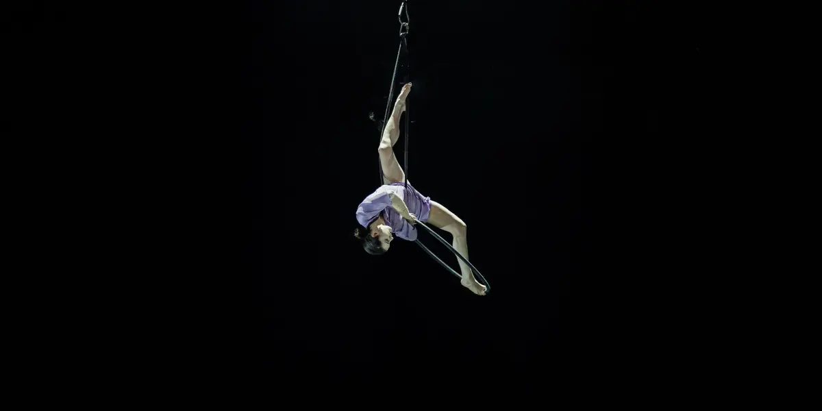 L'artiste aérienne Katarina Sobinkovicova, en justaucorps lavande, exécute une pose acrobatique sur un trapèze, suspendue à l'envers par un harnais sur un fond noir.