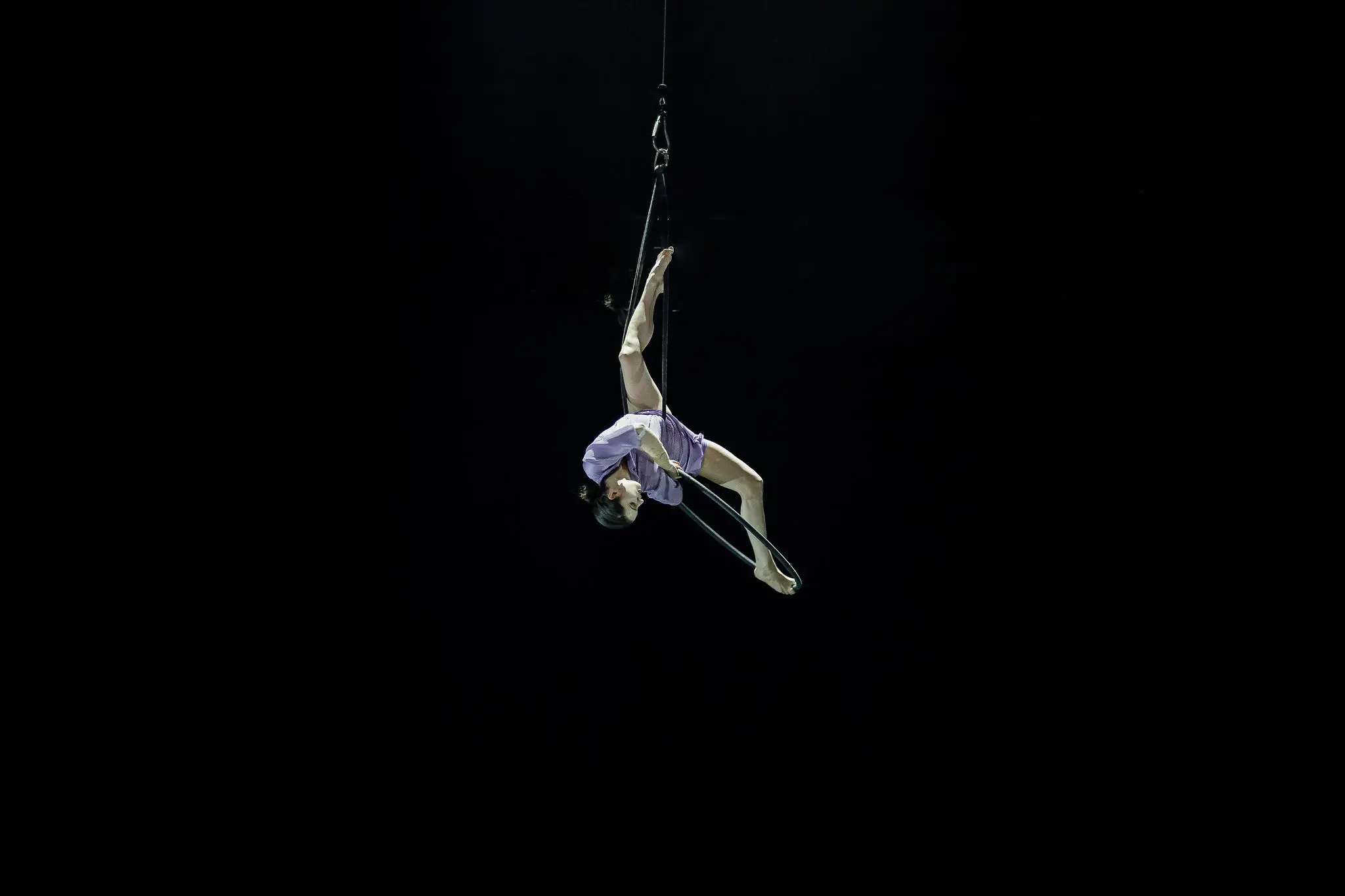 L'artiste aérienne Katarina Sobinkovicova, en justaucorps lavande, exécute une pose acrobatique sur un trapèze, suspendue à l'envers par un harnais sur un fond noir.