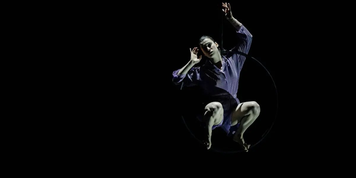 Katarina Sobinkovicova, vêtue d'une chemise violette et d'un short noir, s'exécute sur un cerceau aérien sur un fond sombre, éclairé par un projecteur.