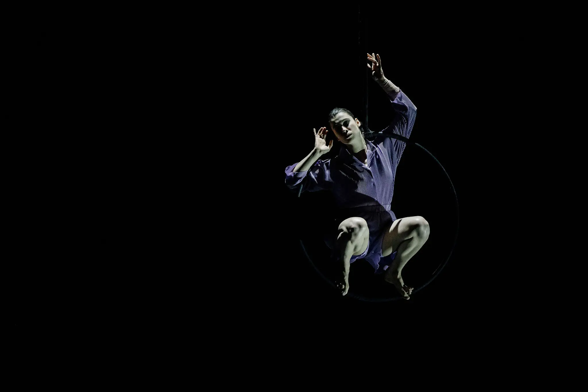 Katarina Sobinkovicova, vêtue d'une chemise violette et d'un short noir, s'exécute sur un cerceau aérien sur un fond sombre, éclairé par un projecteur.