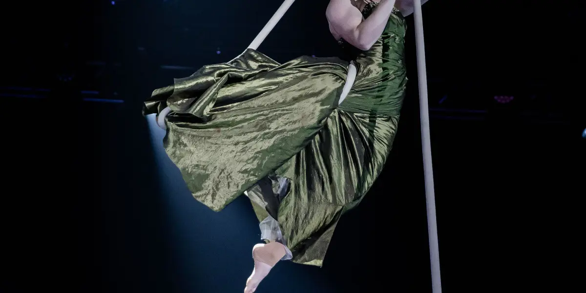 Une artiste aérienne, Flavie Beaulieu, vêtue d'une longue robe verte, se suspend gracieusement à une corde, en plein vol, sur une scène sombre éclairée par un projecteur.