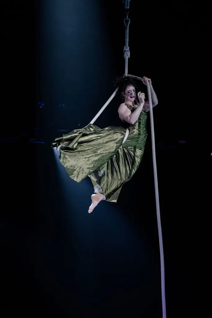 Une artiste aérienne, Flavie Beaulieu, vêtue d'une longue robe verte, se suspend gracieusement à une corde, en plein vol, sur une scène sombre éclairée par un projecteur.