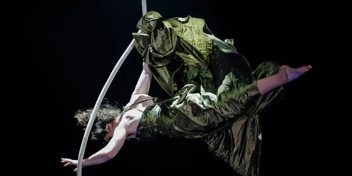 L'artiste aérienne Flavie Beaulieu, vêtue d'une robe verte fluide, est suspendue à une corde blanche, dans les airs, sur une scène sombre, avec des projecteurs spectaculaires en arrière-plan.