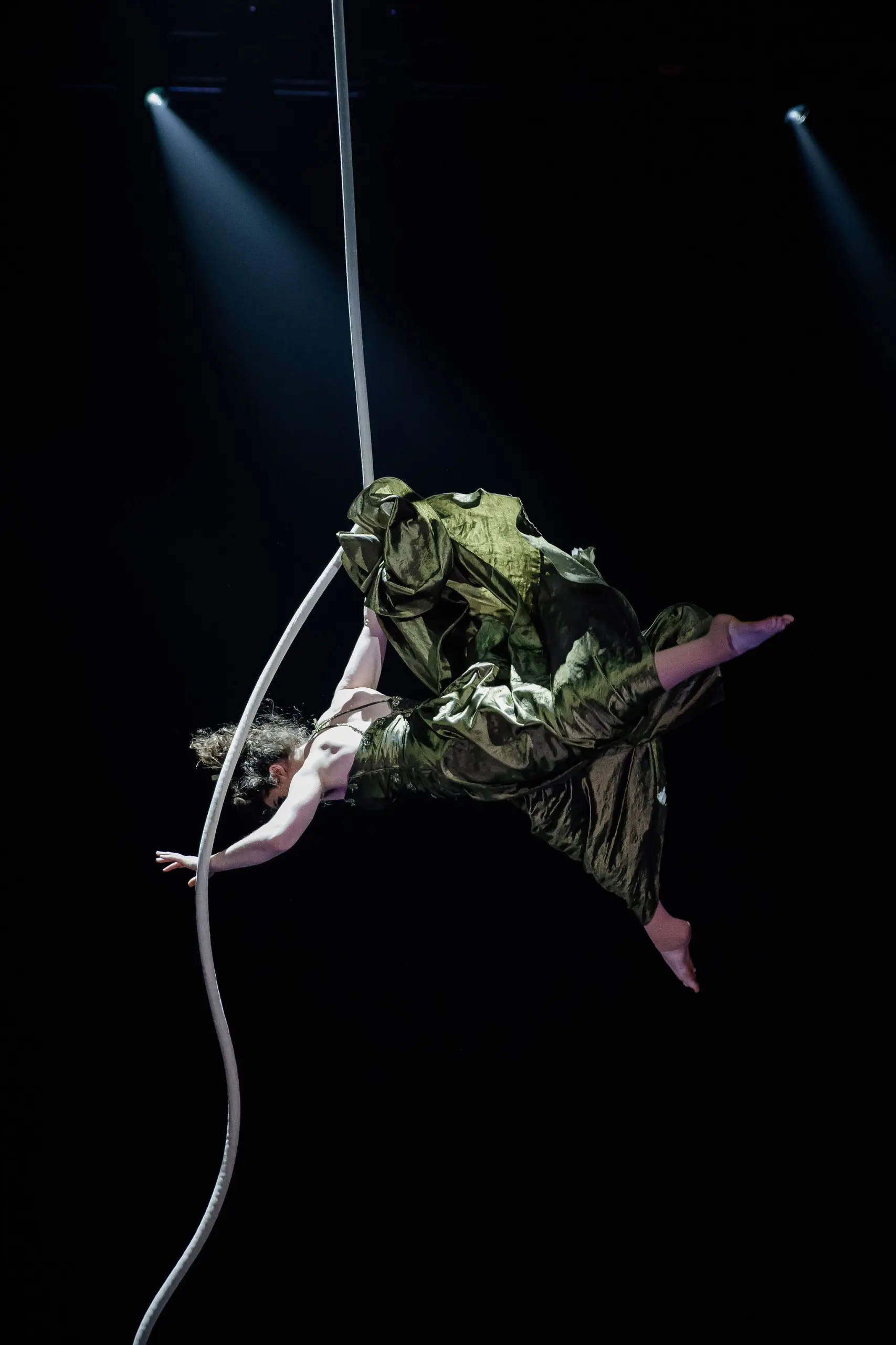 L'artiste aérienne Flavie Beaulieu, vêtue d'une robe verte fluide, est suspendue à une corde blanche, dans les airs, sur une scène sombre, avec des projecteurs spectaculaires en arrière-plan.