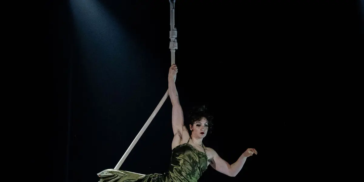 Flavie Beaulieu, en robe verte, effectue un numéro d'acrobatie aérienne, suspendue à une corde accrochée au plafond, avec des lumières de scène en arrière-plan dans un décor sombre.