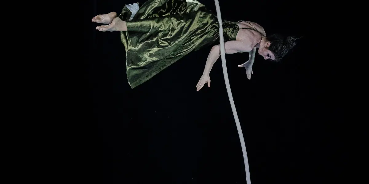 L'artiste aérienne Flavie Beaulieu, vêtue d'une robe verte, est suspendue à une corde blanche, la tête en bas, sur un fond sombre, éclairée par un projecteur placé en haut.