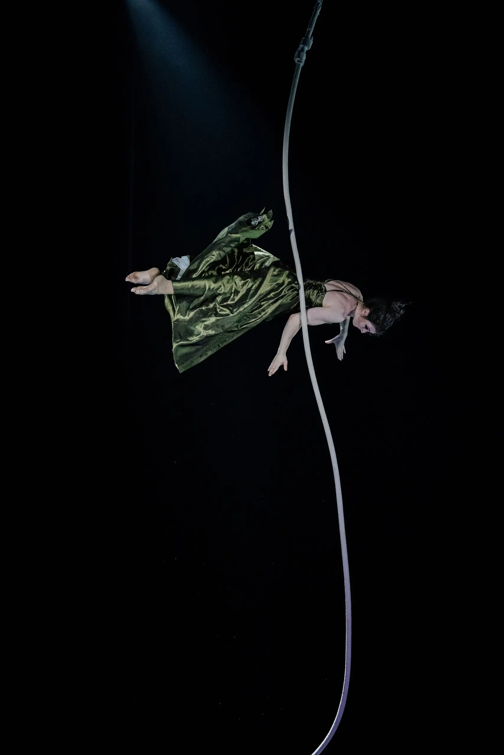 L'artiste aérienne Flavie Beaulieu, vêtue d'une robe verte, est suspendue à une corde blanche, la tête en bas, sur un fond sombre, éclairée par un projecteur placé en haut.